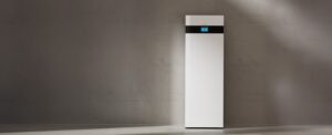 Tall white, minimalist refrigerator standing against a gray concrete wall with a subtle spotlight on it in an industrial setting.